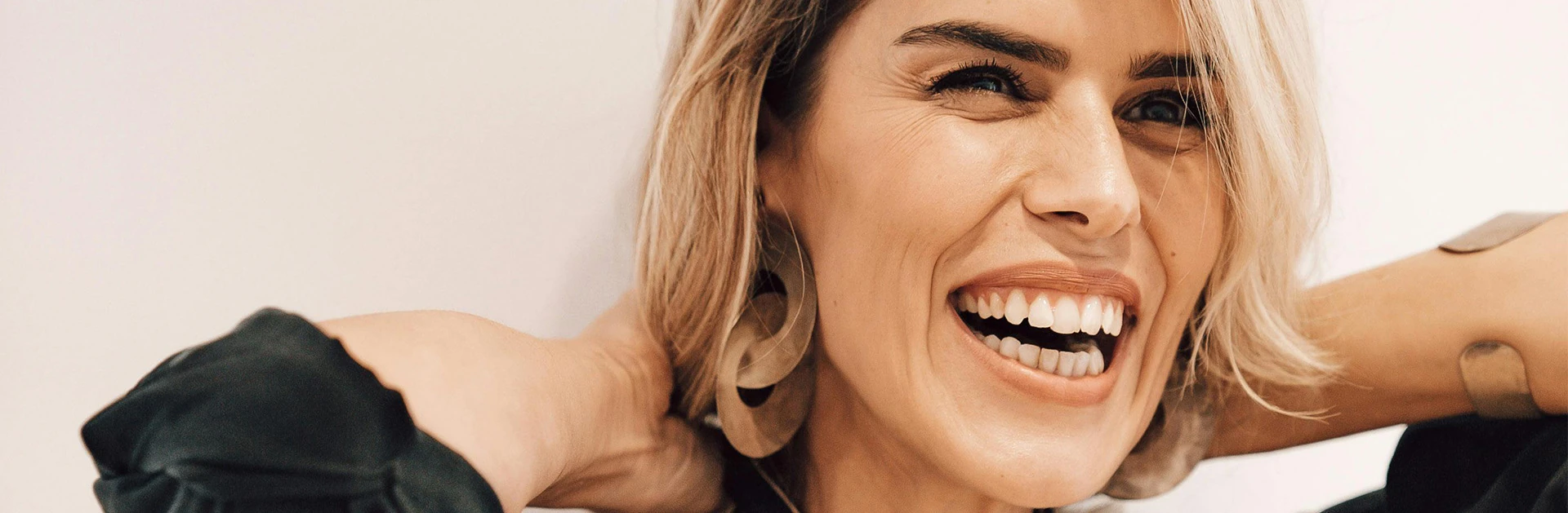 Lächelnde Frau mit blonden Haaren und großen Ohrringen, die ihre Haare zurückzieht und eine entspannte Haltung zeigt.