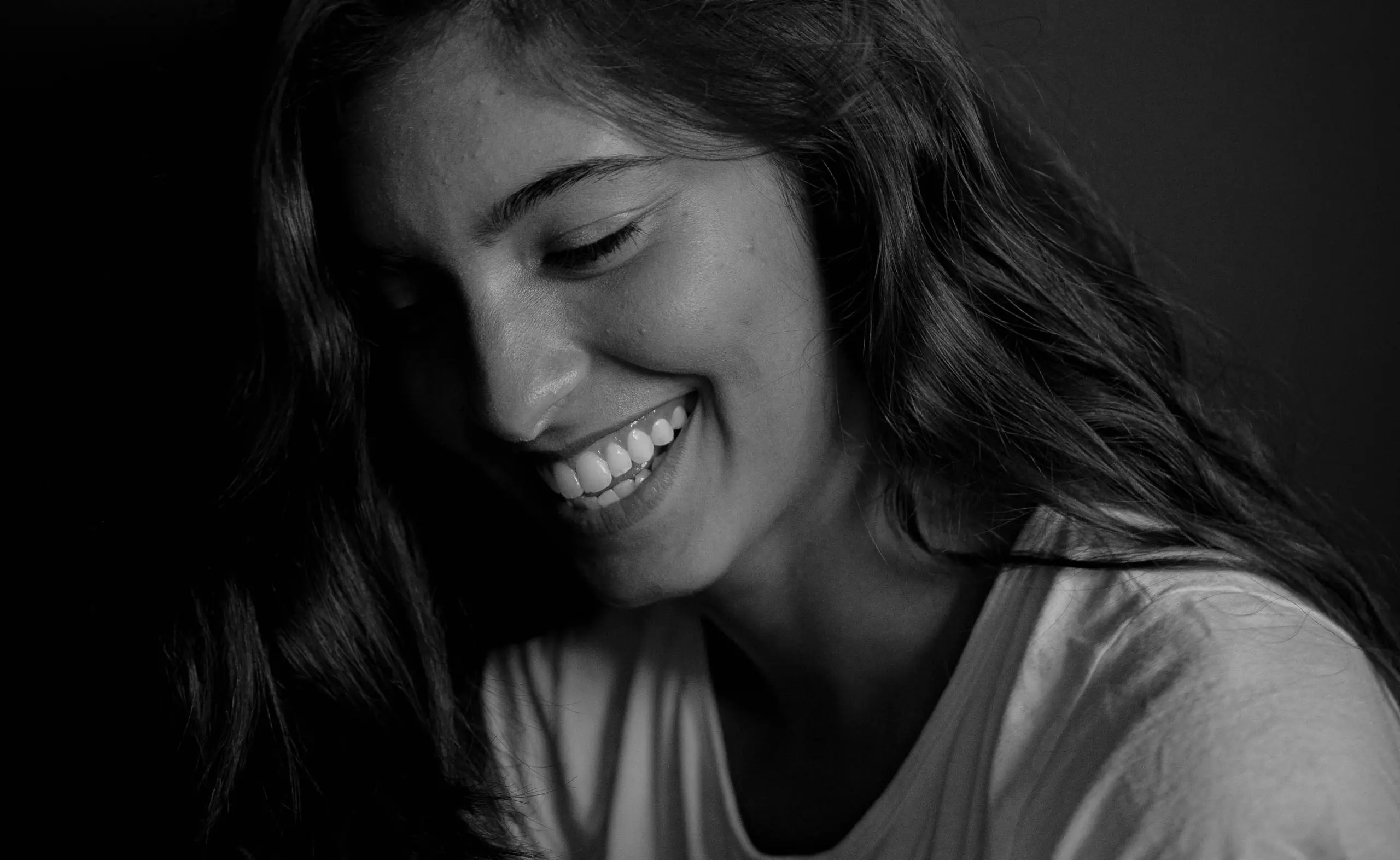 Lächelnde Frau mit langen, lockigen Haaren, die in einem hellen T-Shirt abgebildet ist. Ihr Gesicht zeigt eine entspannte und freundliche Ausstrahlung.