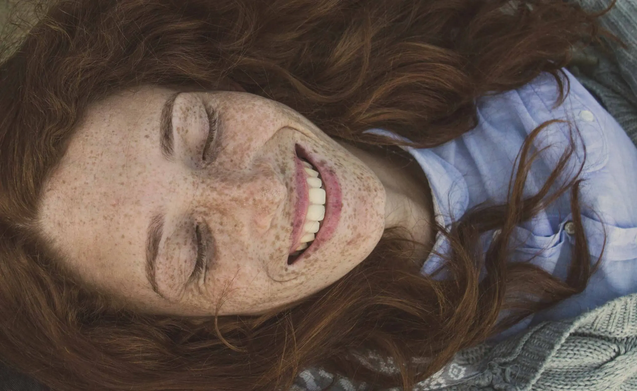 Lächelnde Frau mit roten Haaren und Sommersprossen, die entspannt auf dem Rücken liegt und die Augen geschlossen hat.