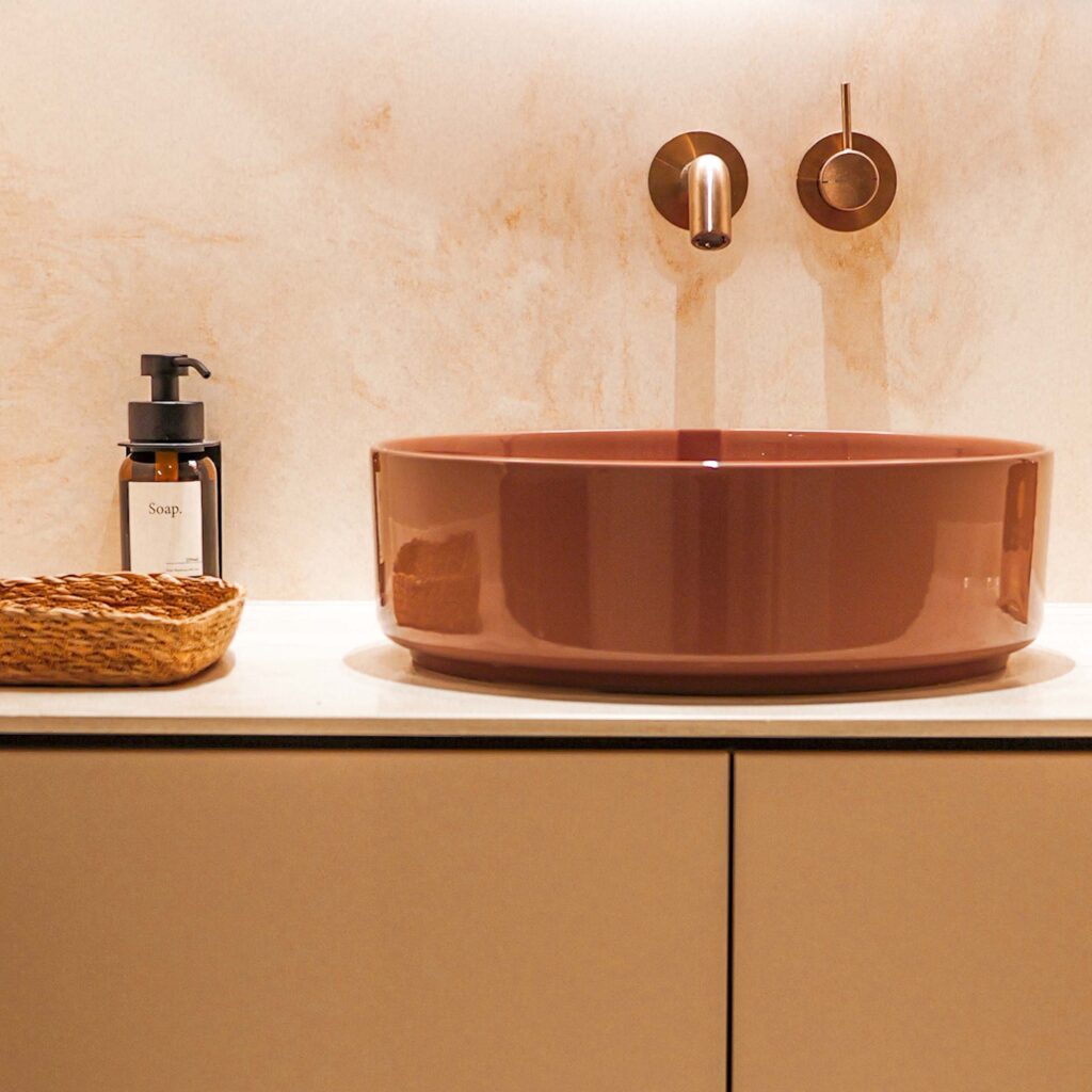 Braune runde Waschschüssel aus Keramik vor heller Arbeitsplatte, dunkler Seifenspender mit Etikett, gewebter Korb und zweifache Wandhaken aus Metall – SP64 Praxisbild