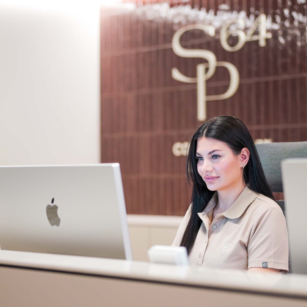 SP64 Praxis Empfang mit moderner Rezeptionistin am Computer vor einer braunen Wand mit Logo-SP64; stilvolles, helles Ambiente, professionelle Atmosphäre