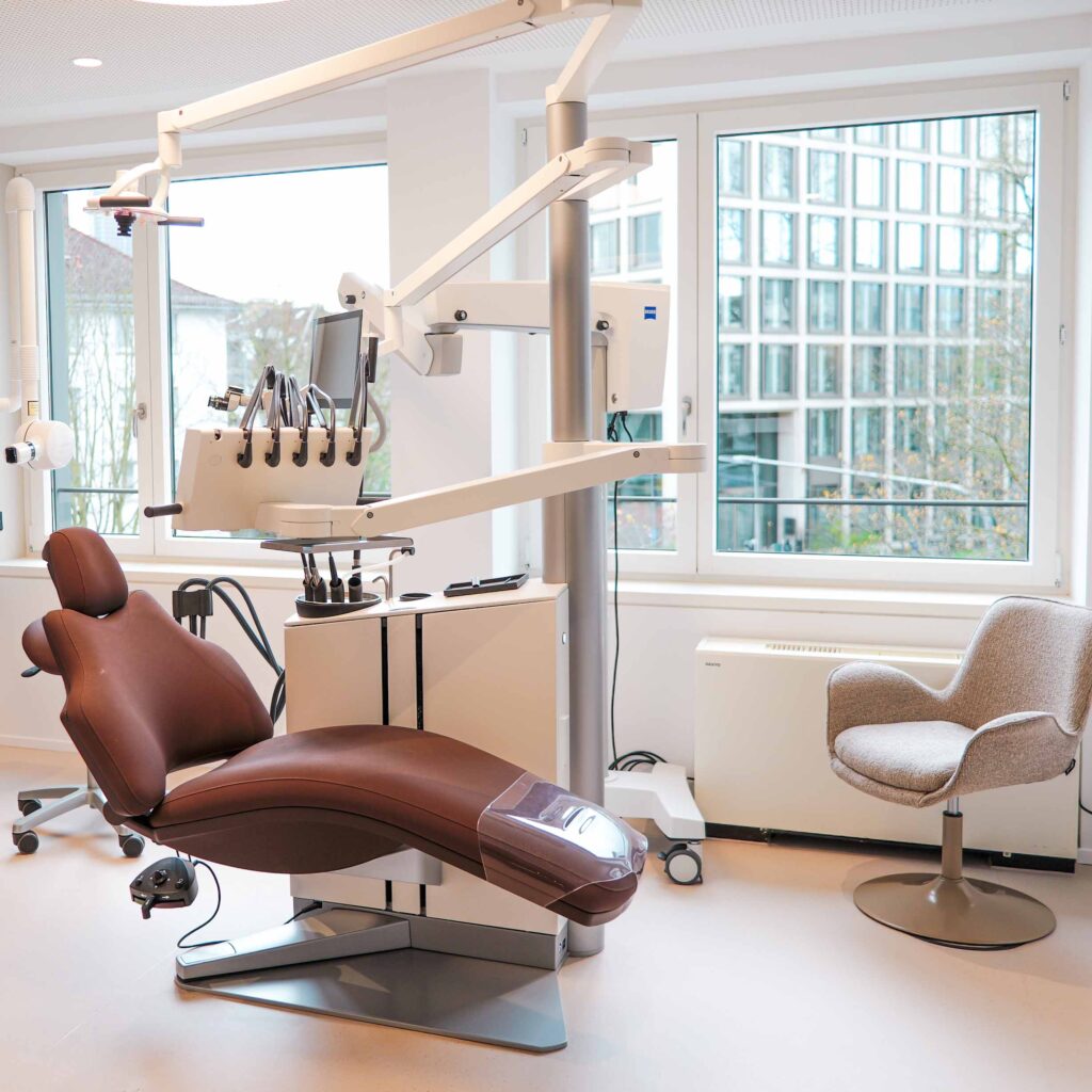 Moderne Zahnarztpraxis-Szene mit braunem Behandlungsstuhl, High-Tech-Behandlungsgerät und großen Fenstern; SP64 Praxisinhaber Stefan Panait, Dr. Jacqueline Ackermann, Dr. Leonidas Bsat