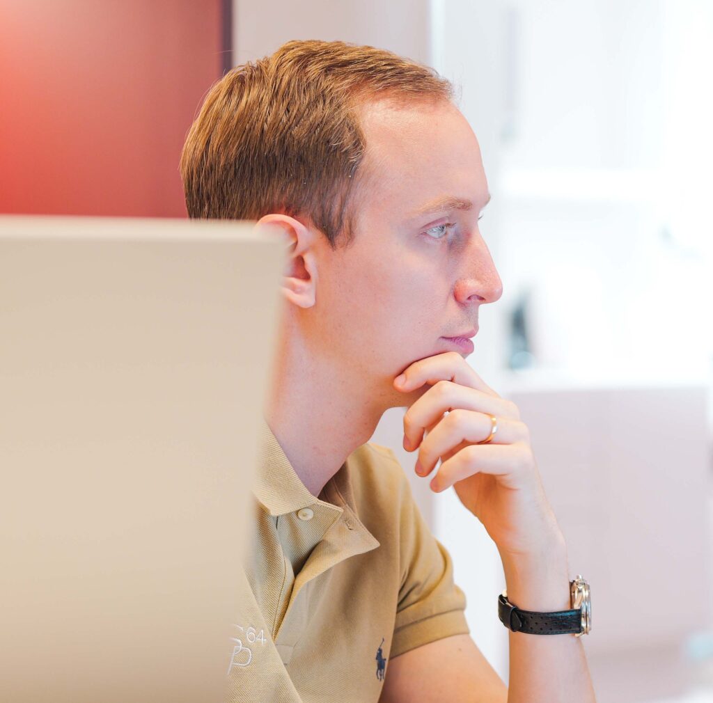 Stefan Panait in einem hellbeigen Polo-Hemd, nachdenklich am Arbeitsplatz, rechts Hintergrund ist hell, linke Hand unterstützt das Kinn, goldene Uhr am linken Handgelenk