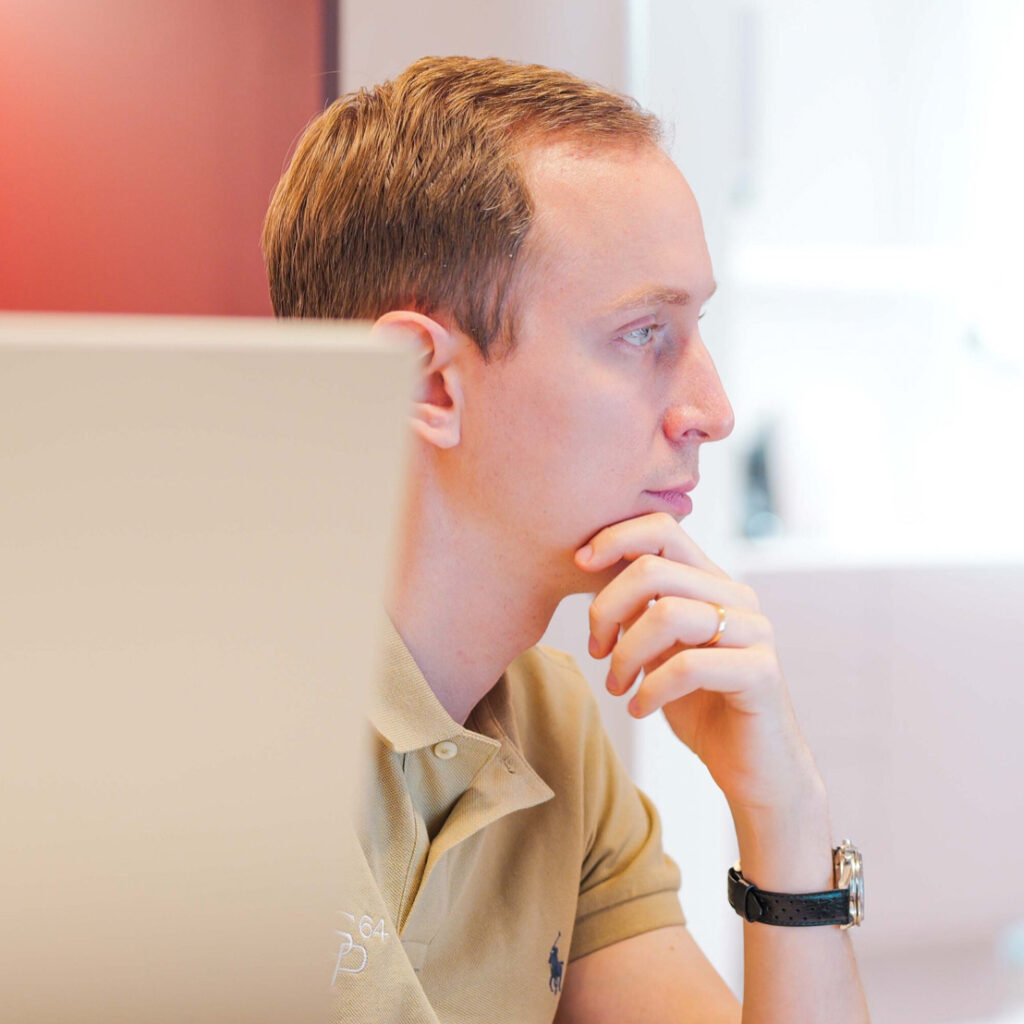 Stefan Panait in einem hellbeigen Polo-Hemd, nachdenklich am Arbeitsplatz, rechts Hintergrund ist hell, linke Hand unterstützt das Kinn, goldene Uhr am linken Handgelenk Stefan_Panait